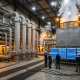 Industrial thermal management facility with steel pipes, heat exchangers, control panels, and temperature monitoring displays with steam rising from equipment.