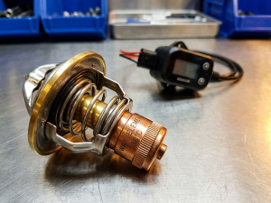 Wax element thermostat with metallic housing and brass components on technical surface with electronic thermostat in background