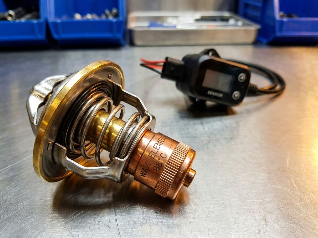 Wax element thermostat with metallic housing and brass components on technical surface with electronic thermostat in background