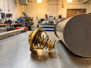 Precision automotive thermostat with brass actuator beside honeycomb catalytic converter on metal workbench in engineering lab