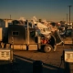 Commercial trucks at fuel station with heat waves rising from engines, open hood revealing glowing components, fuel dispensers showing high costs in golden hour light