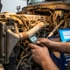 Fleet manager's hands holding digital thermometer over truck engine bay with cooling components and thermal sensors