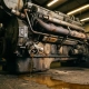 Cracked diesel engine thermostat housing leaking coolant onto concrete floor in industrial maintenance bay