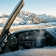 Precision car engine under open hood on a mountain road, snow-capped peaks in background, golden late-afternoon light across metal surfaces.