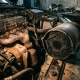 Clogged catalytic converter with heavy carbon buildup and blocked honeycomb core beside an engine bay with exhaust-stained components.