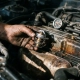 Mechanic's grease-stained hand holding a car thermostat near an engine bay with coolant hoses visible in the background.