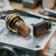 Disassembled mechanical thermostat with coiled wax element beside an electronic thermal sensor on a machined aluminum engine component with metal shavings.
