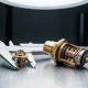 Precision thermostat component held in caliper jaws beside a larger thermostat valve, showing brass and copper internal mechanisms on a steel workbench.
