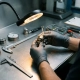 Gloved technician inspecting a precision thermostat component under amber inspection lamp on steel workbench with calibration gauges nearby.