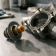 Cross-sectioned automotive thermostat exposing its waxy cylindrical element, placed beside a disassembled engine coolant housing on a steel workshop surface.