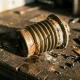Corroded brass thermostat bellows with stress fractures and accordion-fold fatigue on a worn metal workshop surface, surrounded by mineral deposits.