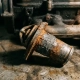 Corroded car engine thermostat valve with rust and exhaust residue on a grimy workshop surface.
