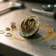 Cracked and corroded wax pellet thermostat on a steel workshop surface with dried coolant residue and small metal components nearby.